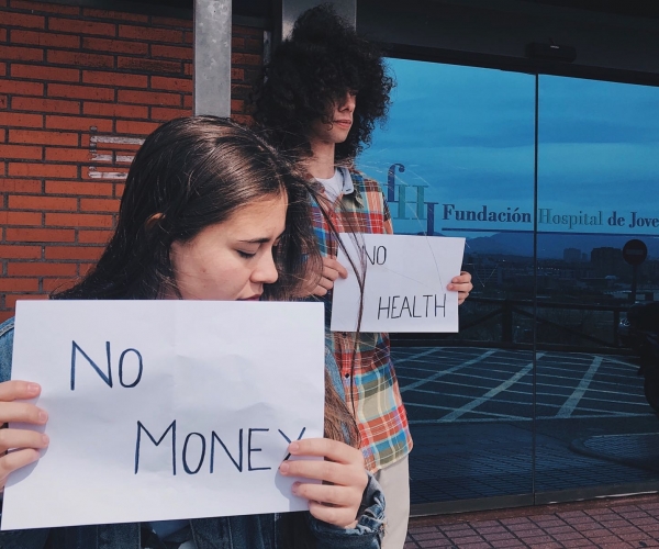 Un hombre y una mujer se manifiestan a la entrada de un hospital a favor de la salud pública con dos pancartas que reflejan un problema mundial: sin dinero, no hay salud. La mujer porta la pancarta con el mensaje “no money” para reflejar la desigualdad salarial entre hombre y mujer.  En muchos países como España existe una diferencia de hasta el 15% en el salario.  Miguel Villa Fernández, 2 de mayo de 2018, Hospital de Jove, Gijón.